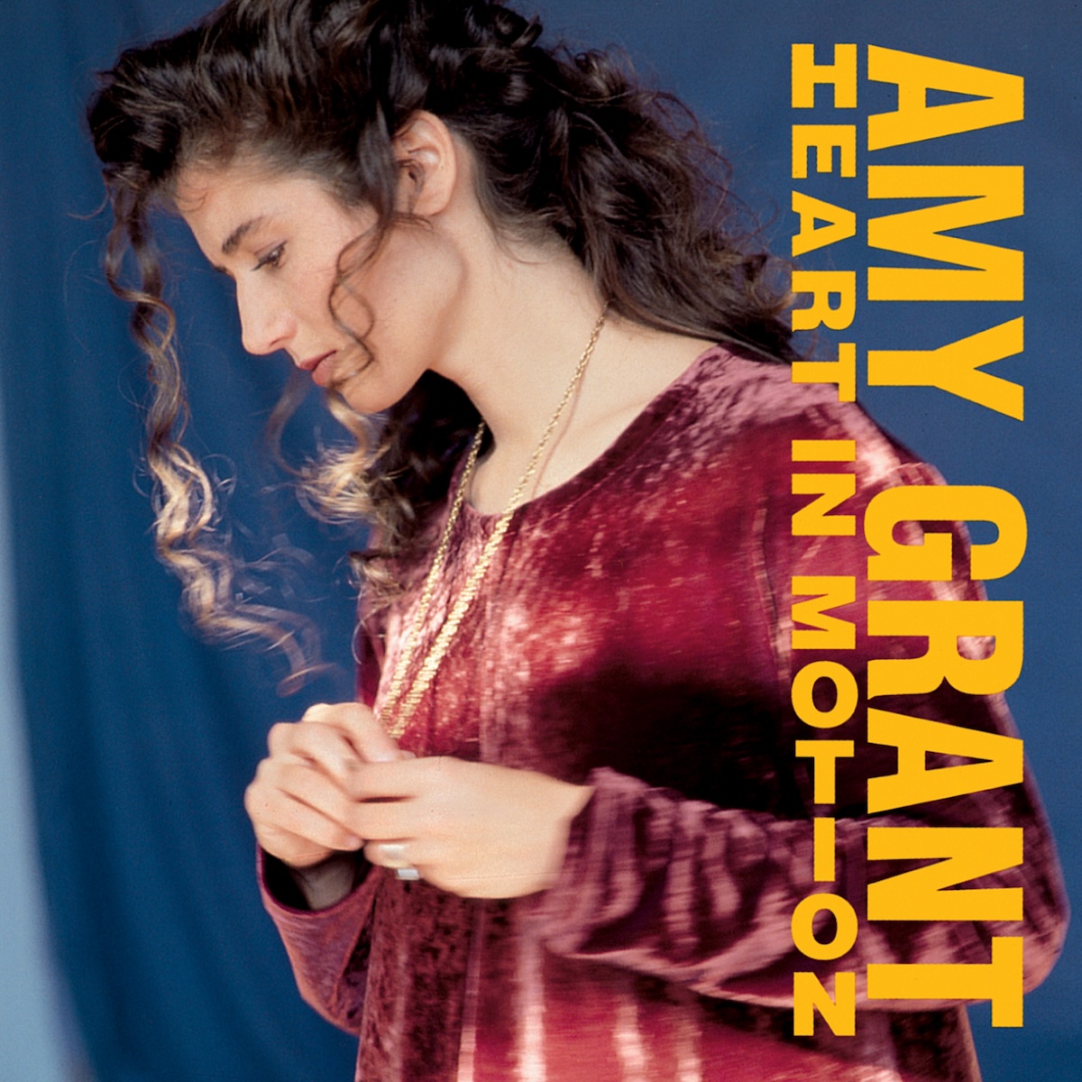 Album Art of Amy Grant's Heart In Motion album - In front a blue backdrop, a young white woman in a red dress is shown in profile and looking down and to the left. She's got a gold chain around her neck and her hands are clasping the item on the chain just in front of her chest. She's got long, brown curly hair. Printed above the photo on the right side is the artist's name down the right side vertically. And printed just to the left of that is the album title in smaller yellow text, with the first letter printed vertically, and the rest of the letters printed horizontally.