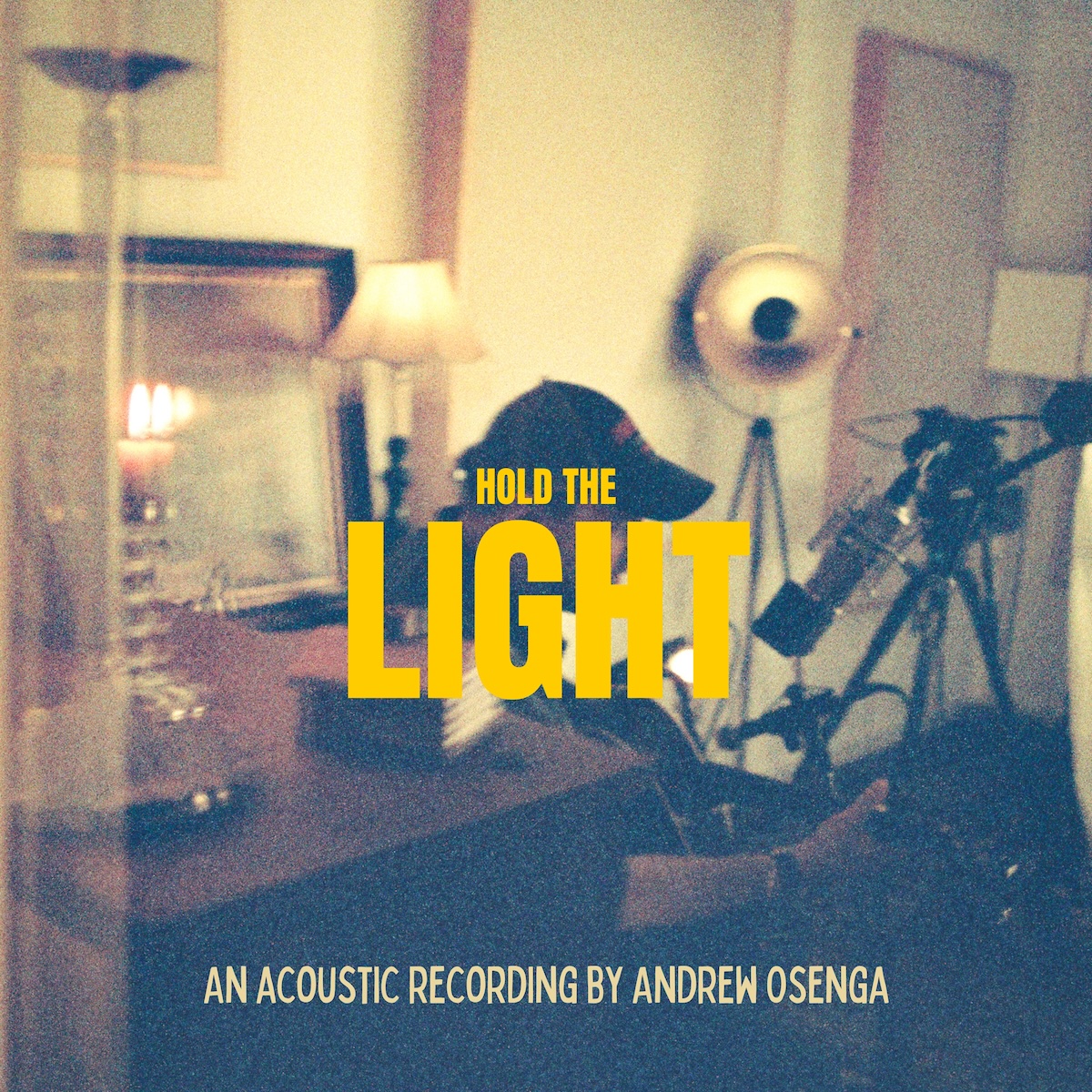 Album Art of Andrew Osenga's Hold The Light album - a grainy color photo of a middle-aged man in a baseball cap playing an acoustic guitar in a small room filled with furniture, lamps and video and audio recording equipment. Above the center of the photo in a bright yellow block text is the words 'Hold The' in small print, and 'Light' below it in much bigger print. At the bottom in a much thinner, lighter yellow print it says ‘An acoustic recording by Andrew Osenga'.