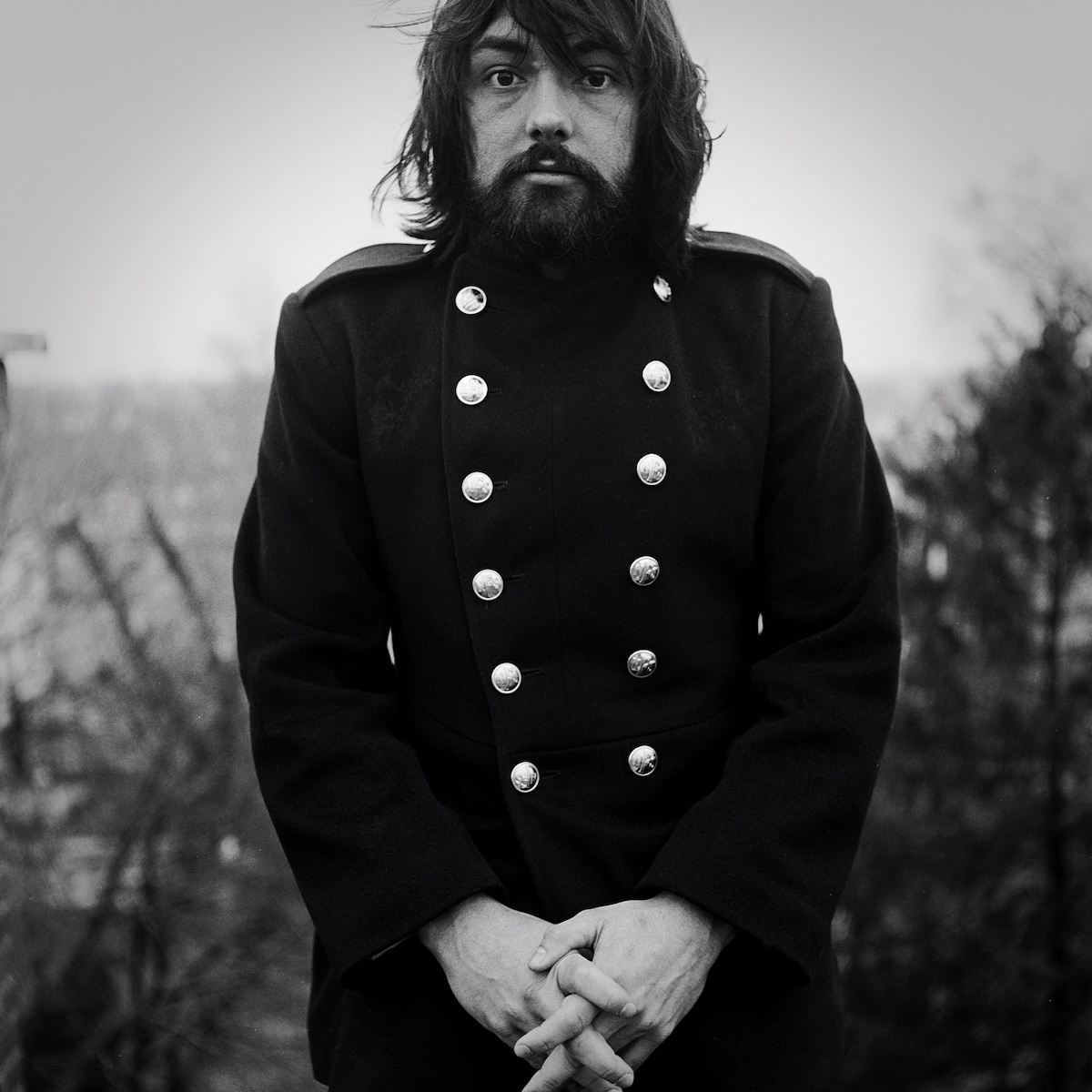 Album Art of Andrew Peterson's A Liturgy, A Legacy, and The Songs Of Rich Mullins album - A black-and-white photo of a man in a funny double-breasted coat that has large silver buttons on both the right and the left of the center. He's got a somewhat surprised look on his bearded face and has thick, dark hair. His hands are clasped together in front of his waist and and looks a bit awkward. The top of his head is not visible nor are his legs, and it looks like the background is a field of some sort.