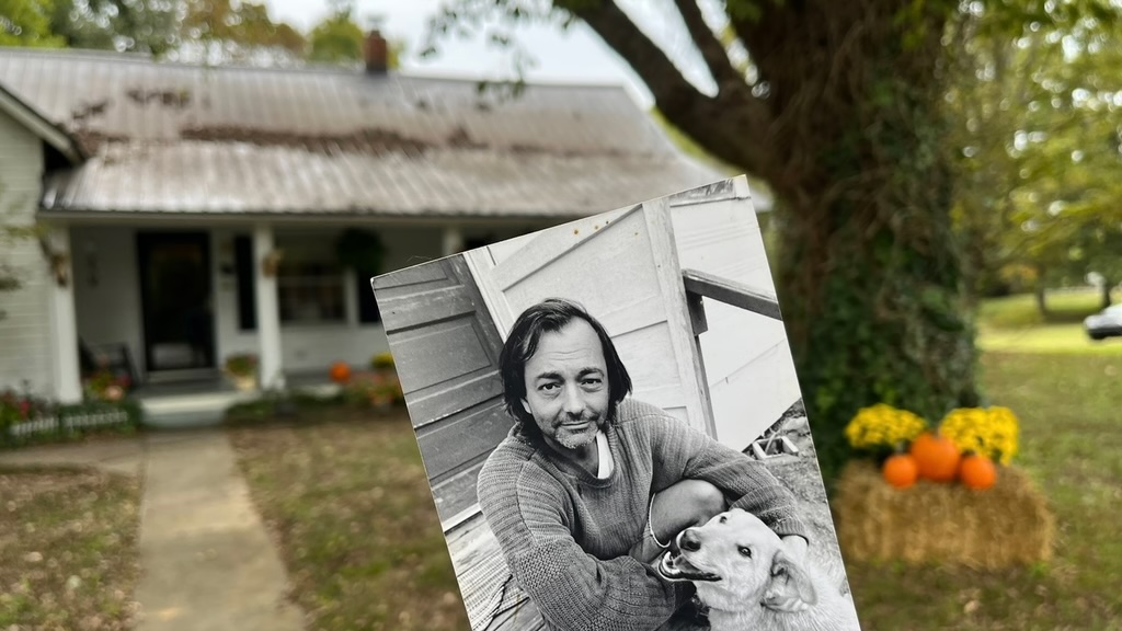 A picture of a small, one-level house with a tree in the front yard photographed in the fall with fallen leaves on the ground. It has a large front porch. In front of that photo is held a black-and-white photo of a young man with a dog sitting on a front step. Click to back the Kickstarter Campaign