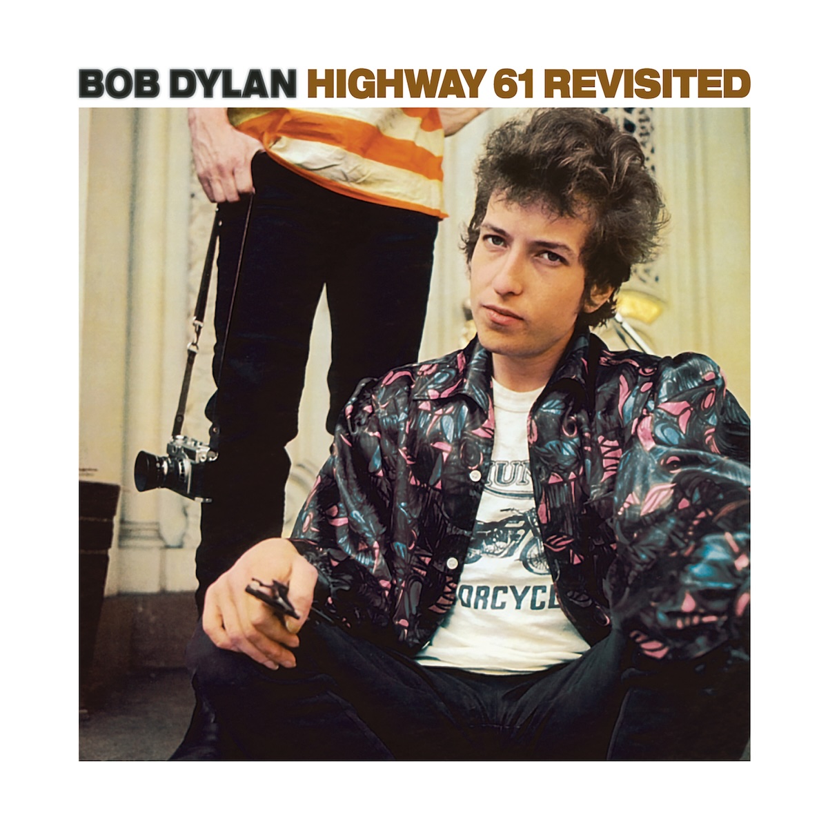 Album Art of Bob Dylan's Highway 61 Revisited album - With a generous white border around it, a color photo of a young white man with curly hair at least a few inches long is looking somewhat annoyedly at the camera. He seems to be sitting down because someone's legs and waist can be seen behind them with black pants and an orange-and-white striped shirt and holding a camera on a strap.</body></html>