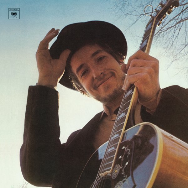 Album Art of Bob Dylan's Nashville Skyline album - A photo of a young white man with a short beard and mustache in a black coat wearing a black hat, and he's holding his hand just below the hat like he's about to tip his hat. He has a noticeable smile and is photographed from below, looking down at the camera while holding his guitar in his left hand in front of him. Behind him can only be seen the blue sky, bluer towards the top and more white near the bottom. In the top right, by the neck of the guitar, the bare branches of a tall tree can be seen.