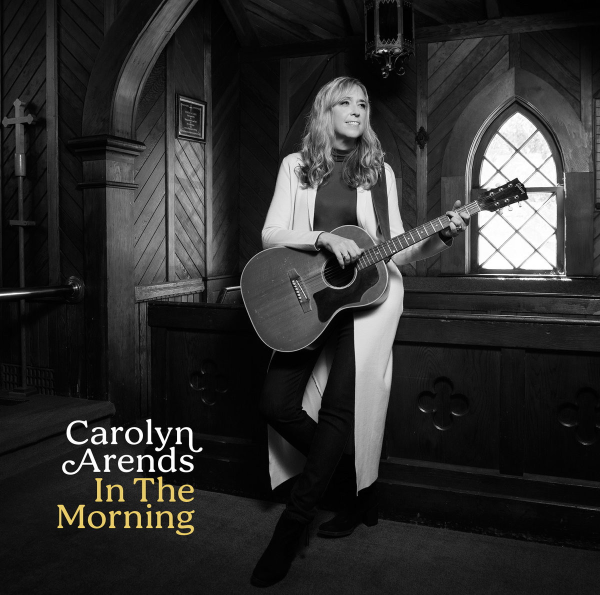 Album Art of Carolyn Arends's In The Morning EP - A black-and-white photo of a white woman standing against a wooden railing and holding an acoustic guitar in some sort of small, rustic chapel with wooden walls and a small, simple glass window. She has blond hair down past her shoulders and is looking off to her left, while she has her hands on the guitar like she might start playing any moment. In the bottom left, printed in white is the artist's name and then the album title in gold below it.