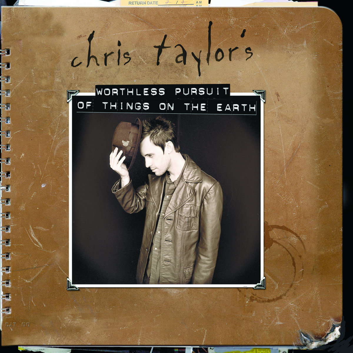 Album Art of Chris Taylor's Worthless Pursuit Of Things On The Earth album - A photo of a wire-bound notebook with a scuffed leather cover that is even missing a bit of the bottom left part. Behind can be seen some pages and other papers sticking out of the top and bottom of the notebook on the very edges of the photo. A few coffee cup stains are also on the leather, just to the bottom right of a photo affixed to the front of the cover. The photo is of a man on a dark background with a brown leather jacket and a brown hat with a short, curved brim. He's holding the hat in his right hand like he is in the process of putting it on. He's slightly smiling and facing mostly to the left of the frame. On the leather above the photo is written in marker or something 'chris taylor's' and then directly on top of the photo is printed 'Worthless Pursuit Of Things On The Earth' by a black label maker.