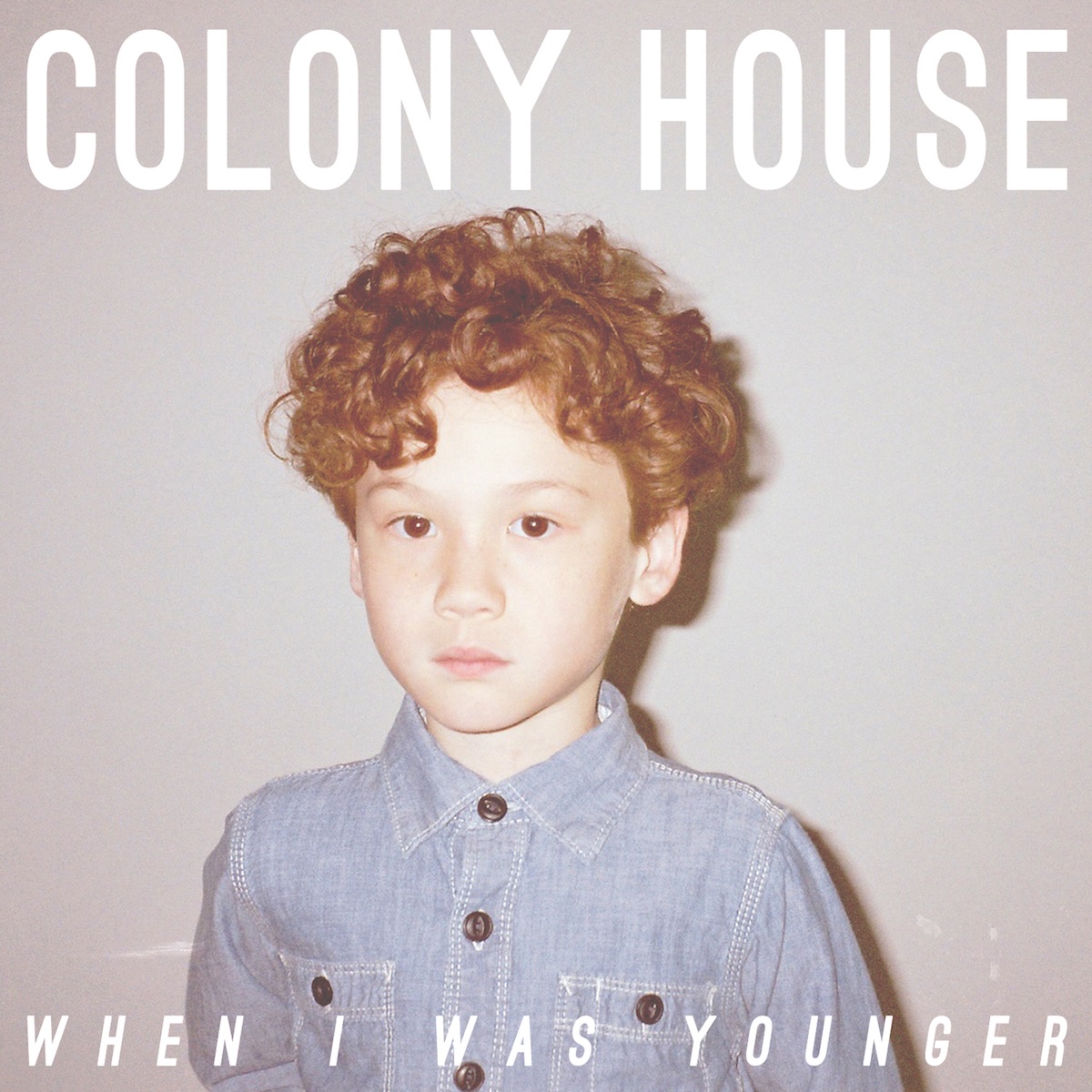 Album Art of Colony House's When I Was Younger album - A photo of a young boy sitting in front of a gray-ish white wall. Only seen from the chest up, he has a denim shirt on and looks fair-skinned with curly, red hair. He looks to be only 3-4 years old and has a fairly blank expression and is looking directly at the camera. At the top, above the boy's head, the band's name is printed in large, white letters. The album title is printed over the photo in smaller white letters across the bottom.