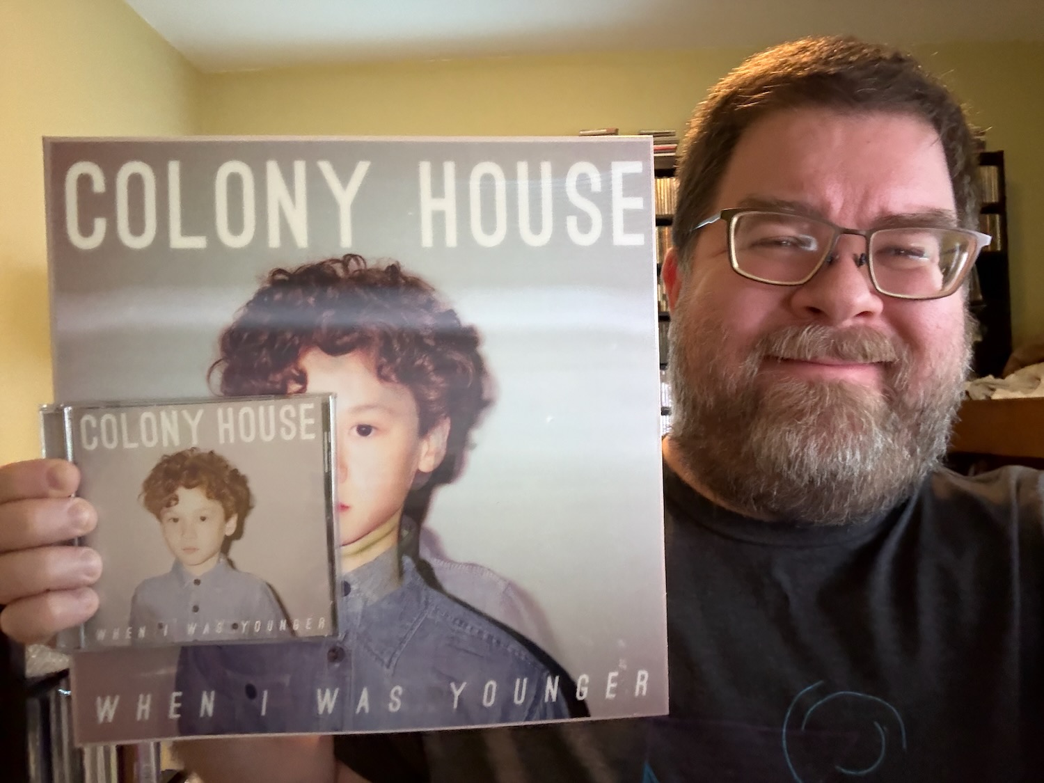 A photo of a middle-aged man with a short beard holding the Colony House LP and the CD in his right hand up towards the camera. On the right, he's smiling and wearing a dark shirt. The room behind him has large racks of CDs, various knick-knacks and and yellow wall.