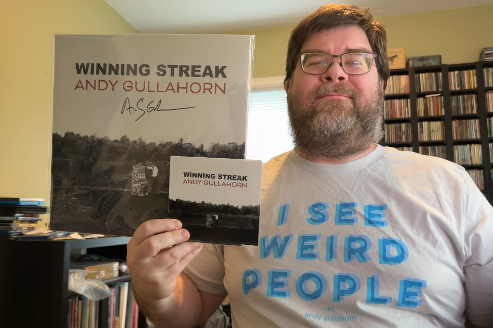 A photo of a middle-aged white man in a home office shelves on the left and back on the right. He was thick glasses, a beard and moustache, and is smiling. He is wearing an off-white shirt that has the words 'I See Weird People' in big letters and 'like Andy Gullahorn' in very small letters below it. In his right hand is a vinyl and CD copy of the Andy Gullahorn Winning Streak album. The vinyl packaging is autographed.