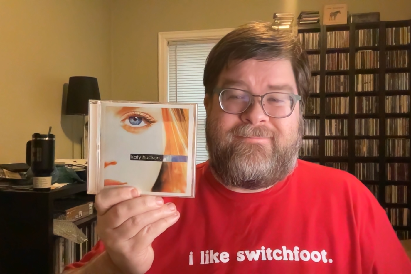 A middle-aged white man photographed in a brightly-colored office room with black CD racks in the background full of CDs, a black lamp on the lower shelf on the left, and a black water tumbler on the left as well. The man is wearing a red T-shirt that says 'I Like Switchfoot' and is holding the Katy Hudson CD in his right hand and smiling.