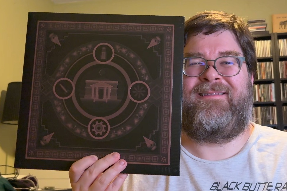 A photo of a man holding the Myst vinyl package up next to his face. He's smiling and looking at the camera with a bit of a toothy smile and is in a yellow-ish room with a lamp and other media behind him.