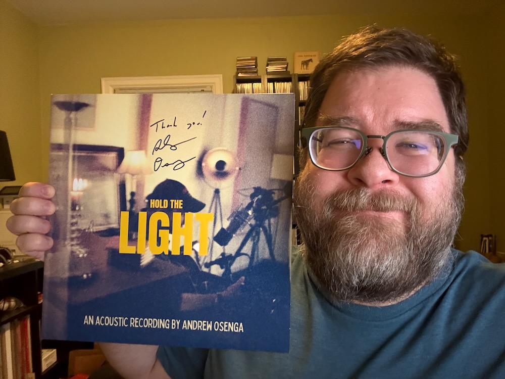 A photo of a smiling man from the shoulders up on the right side holding a vsigned vinyl record to the left. Behind him can be seen CDs and vinyl on shelves. He’s wearing a light blue shirt, has a full beard and mustache and has short, brown hair.