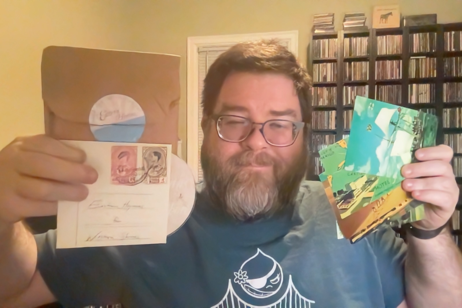 Photo of a rotund man with short brown hair and long, lighter beard and mustache holding a handful of postcards in one hand and a CD in a cardboard slipcase and a brown paper bag in the other hand. He's smiling, has glasses, and has a bunch of CDs in large shelves behind him in a room lit with a golden light.