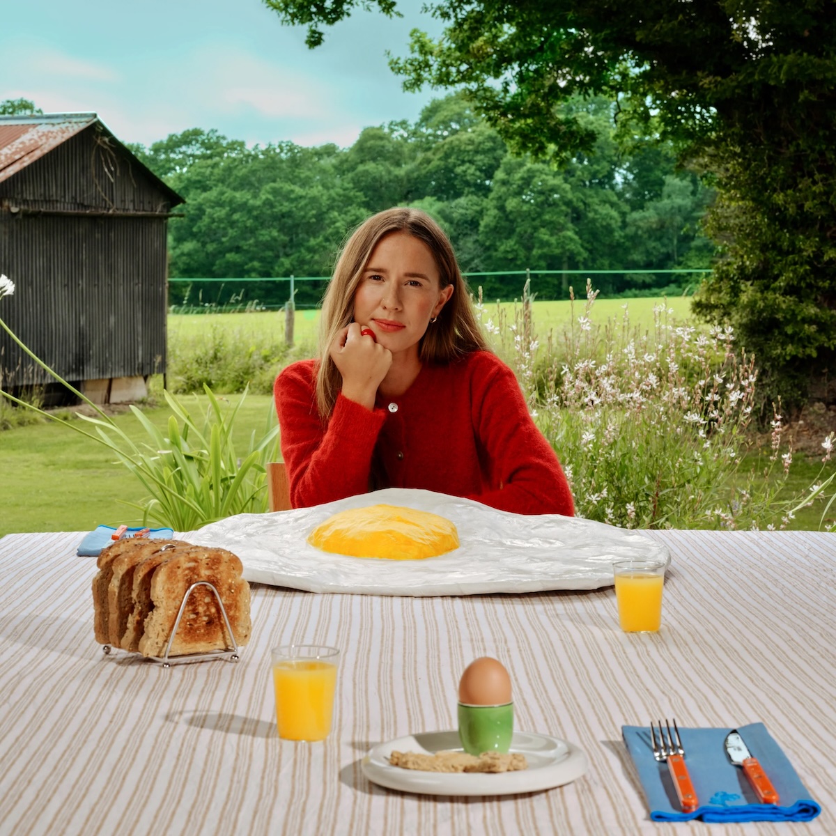 Album Art of Elle Limebear's Welcome To The Bloodline album - A brightly colored photo of a woman sitting at a large picnic table outdoors. The table looks to be in a yard with a tree on the right and an old shed of some sort on the left, plus a fence running between those, and in the far background, past a field, is a line of forest with blue sky above.</body></html>