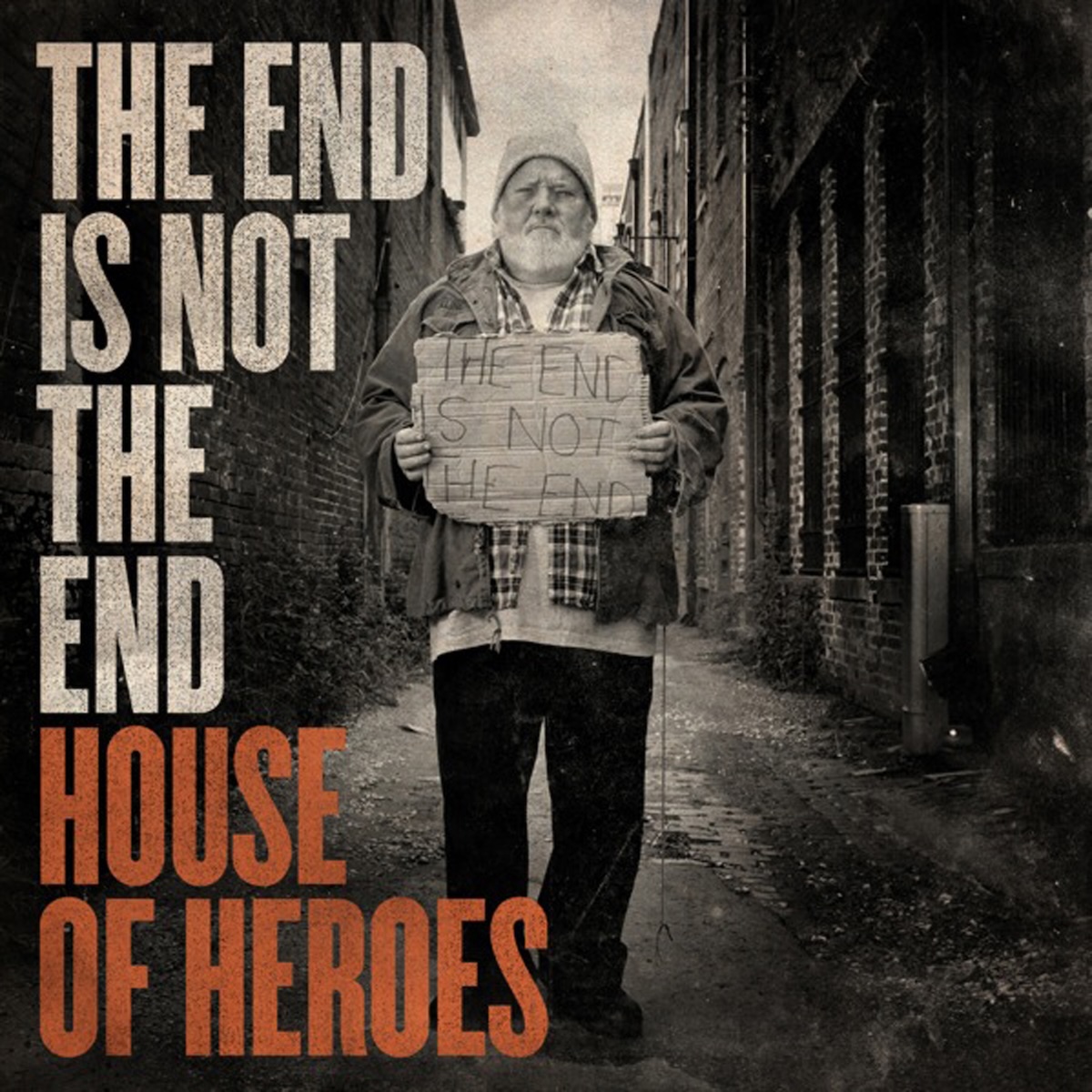 Album Art of House Of Heroes's The End Is Not The End album - In a narrow road between abandoned brick buildings, a black-and-white photo of a man with a white beard and a light-colored beanie is wearing a coat and a few more layers beneath it. In front of his chest he holds a piece of cardboard with the album title, 'The End Is Not The End', written on it in some sort of thin, black handwriting. Down the left side, in large block letters, is also printed the album title in grey with the band's name in the last few lines in orange.