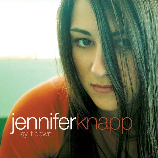 Album Art of Jennifer Knapp's Lay It Down album - A photo of a young woman taken pretty close, where we can see her right shoulder, her face, and some bright sunlight over her shoulder. Her long, brown hair is partially covering her face on the right side of the photo, and covering her shirt that seems red on her shoulders and more gray in the front. her expression is fairly neutral with only a possible hint of a smile. Below her face and to the left is her name, first name in white and last name in red, and then the album title in smaller, light gray below. All of the text is in lowercase and a very thin sans-serif font.