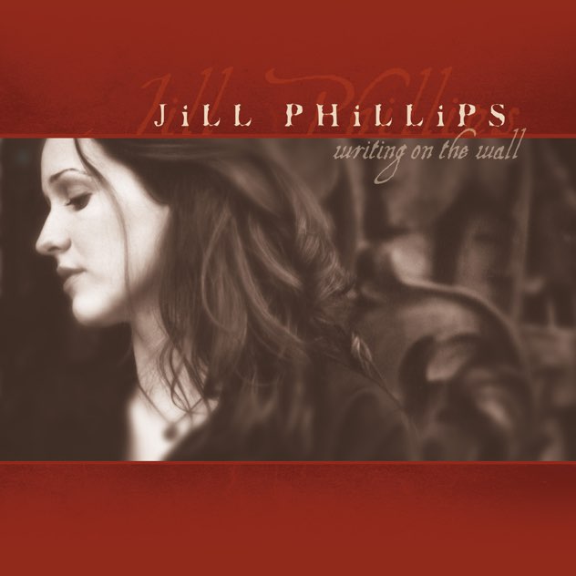 Album Art of Jill Phillips's Writing On The Wall album - On a red background on top and bottom, in the center is a black and white photo of a young, white woman on the left, looking off to the left and slightly down. Only showing her neck and face, and the long, dark hair flowing over her shoulders mixes with a dark, swirling background off to her right. The dark background has some shapes to it, but I can't tell what it's supposed to be. above the picture in a faint, script-like font is the album title, and above the photo on the red background is the the artist name in white, thicker letters with a faint script version underneath it.