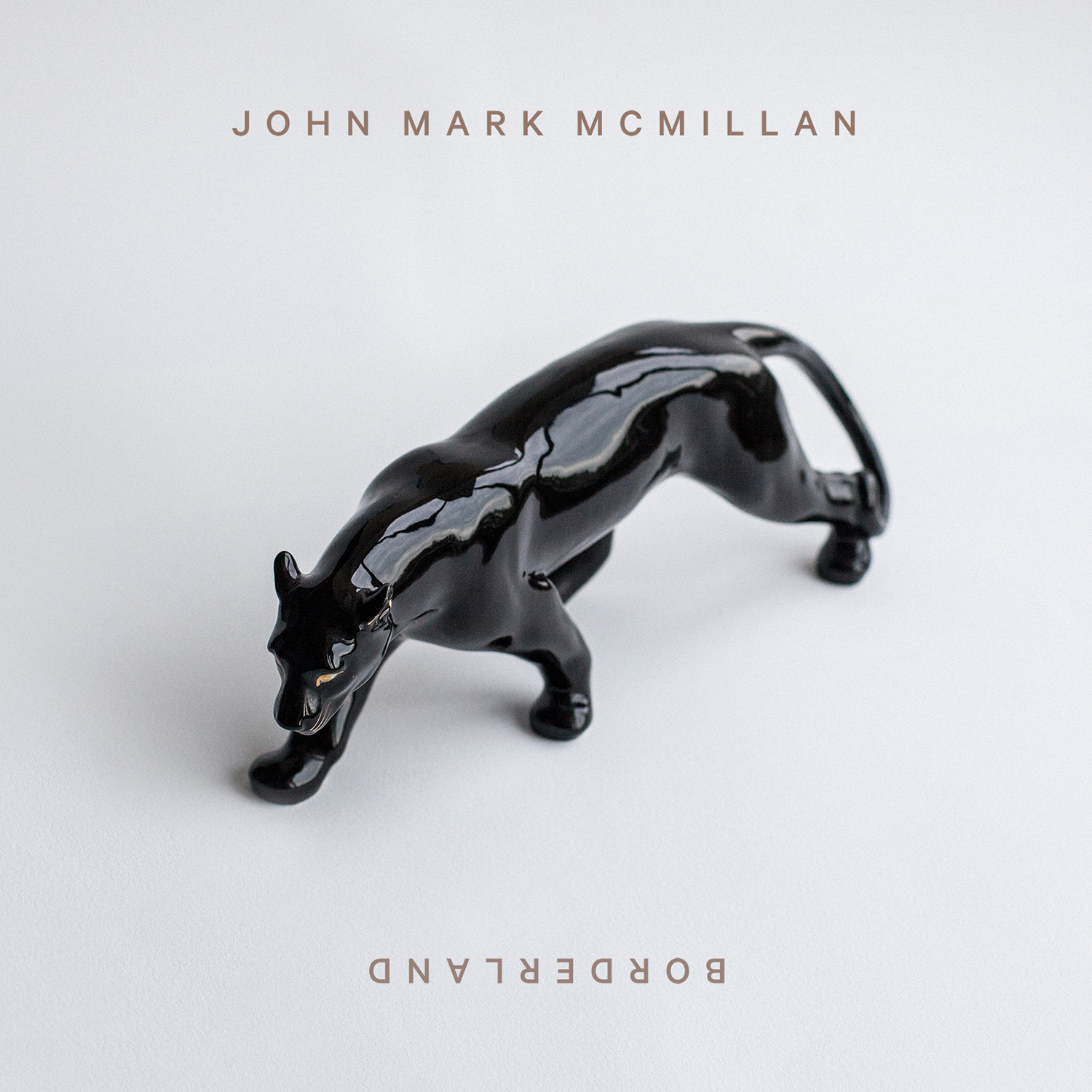 Album Art of John Mark McMillan's Borderland album - A photo of a black panther ceramic statue on a white background, with only a very soft shadow below it and to the left of it. At the top, centered in small golden letters is the artist's name, and on the bottom in gold letters rotated 180 degrees is the album name in the same lettering.