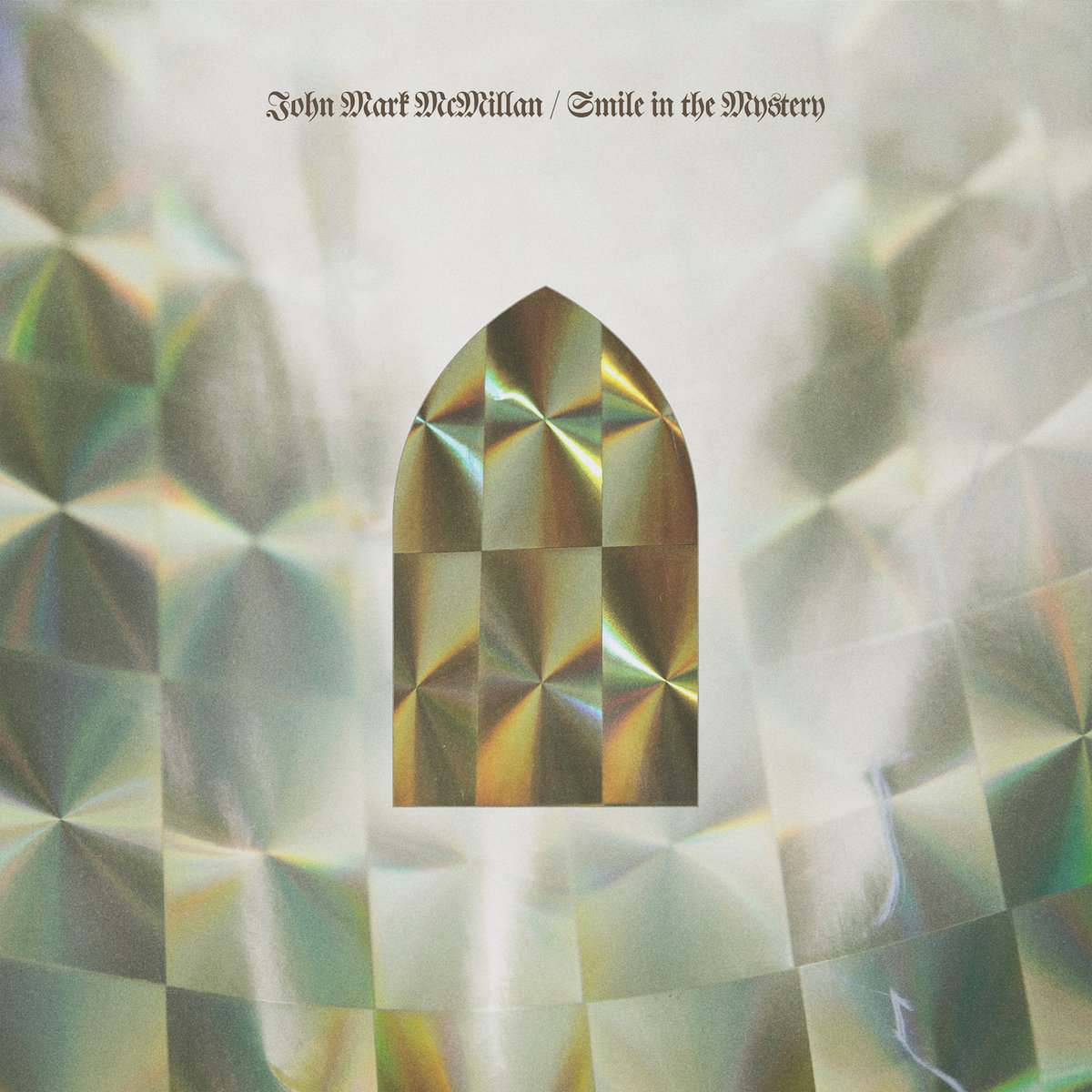 Album Art of John Mark McMillan's Smile In The Mystery album - The background looks like a bunch of shiny paper slightly curved in a circle and with a bit of depth to it. In the middle is a darker shiny, glimmering paper that is in the shape of a stained glass window in a church with a pointy top. At the top, in a classic calligraphy font that's pretty small, it has the artist's name and the album title all on one line in a dark grey.