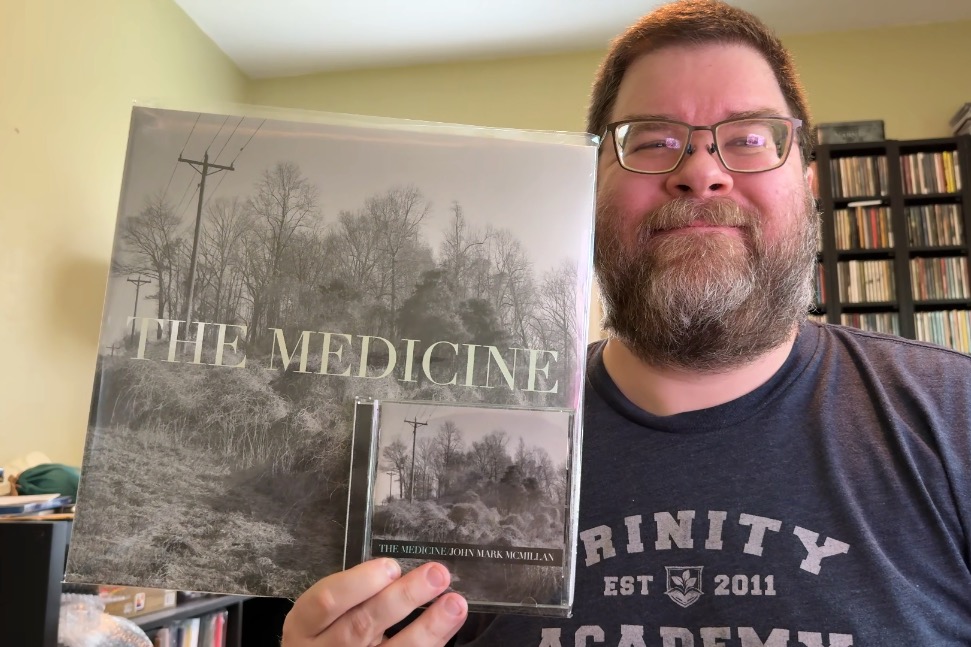 A photo of a middle-aged, smiling man with short hair and a short beard from the chest up in a dark grey T-shirt holding in his right hand 'The Medicine' vinyl with a shiny silver lettering and the CD version as well. Behind him is a room with light green walls and a large set of shelves with lots of CDs.