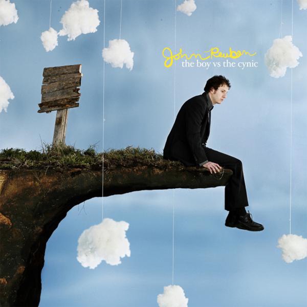 Album Art of John Reuben's The Boy Vs. The Cynic album - A photo of a white man with dark, curly hair sitting on the edge of a precipice and looking down with a bit of a blank stare. The precipice looks like it's a prop set up in a studio, as the dirt/stone of it sticks out comically far to where the person is sitting, like it was designed by a cartoonist. There's also a blank wooden sign next to the precipice but without any text on it. Behind it is a blue background, with white fluffy clouds suspended in front of it, with thin white strings showing that the clouds are hanging from above Above the person's head is the artist's name in a yellow, handwritten script, and the album title in smaller white text below that.