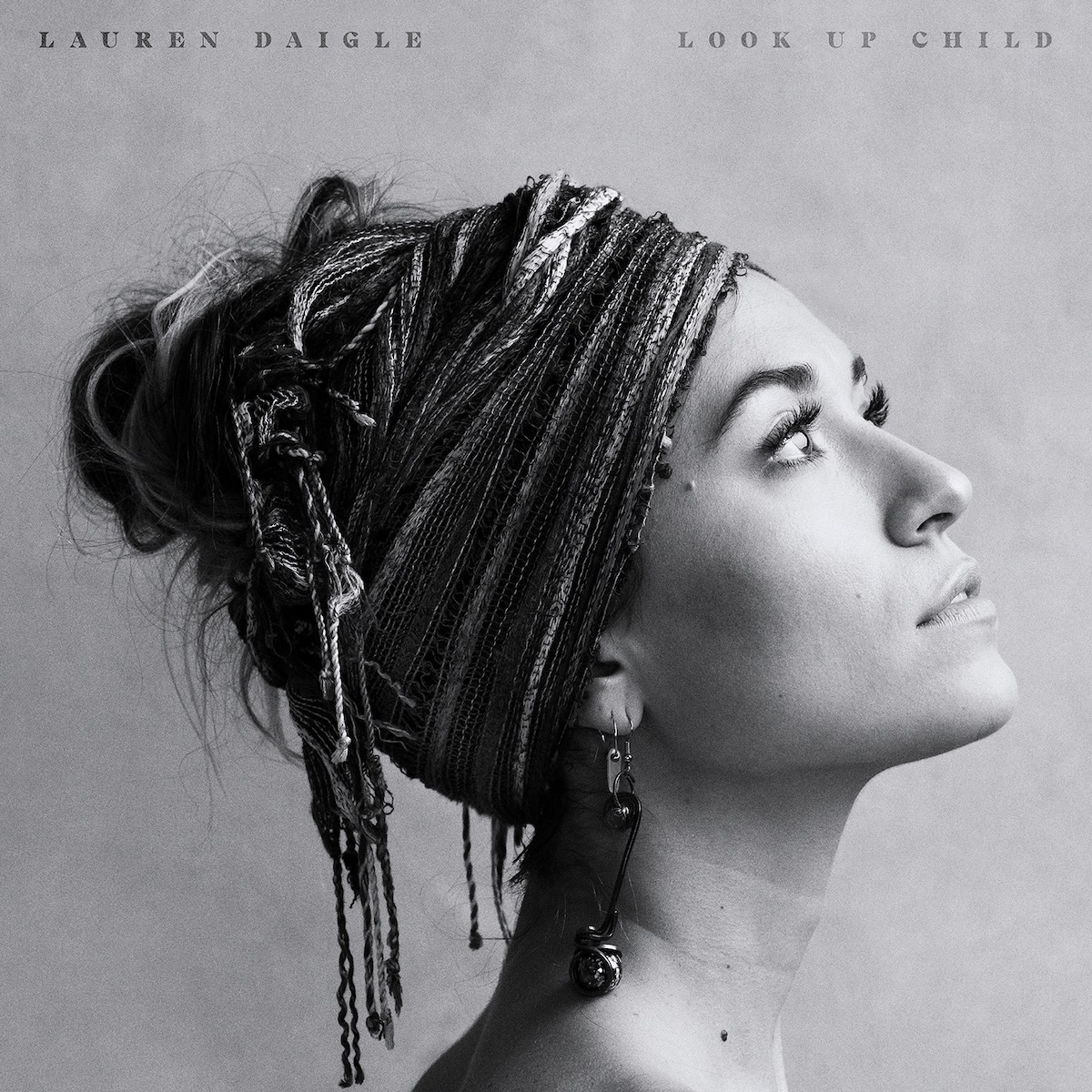 Album Art of Lauren Daigle's Look Up Child album - On a mottled gray background, a black and white photo of a woman from the shoulders up looking to the right an slightly up. The photo is mostly a profile angle, with just a bit of her left eyelash showing behind her nose. Her hair is up inside a cloth that is wrapped around the top and back of her head, covering most of her ears as well. What is visible from her ear has a long earring hanging from it, and her hair covering also has some braided strings hanging from it. At the very top, on the left in a gray-black is the artist name in small print, and on the top right is the album title.