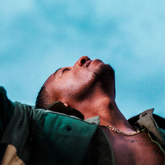 Album Art of Lecrae's Restoration album - Photographed from below, it's a photo of a black man with a green jacket and a bare chest standing in the sunlight and looking up towards the blue sky. His head is slightly back and it looks like he's savoring the moment. He's got a bit of facial hair and stubble and short, dark hair, plus a gold chain around his neck.