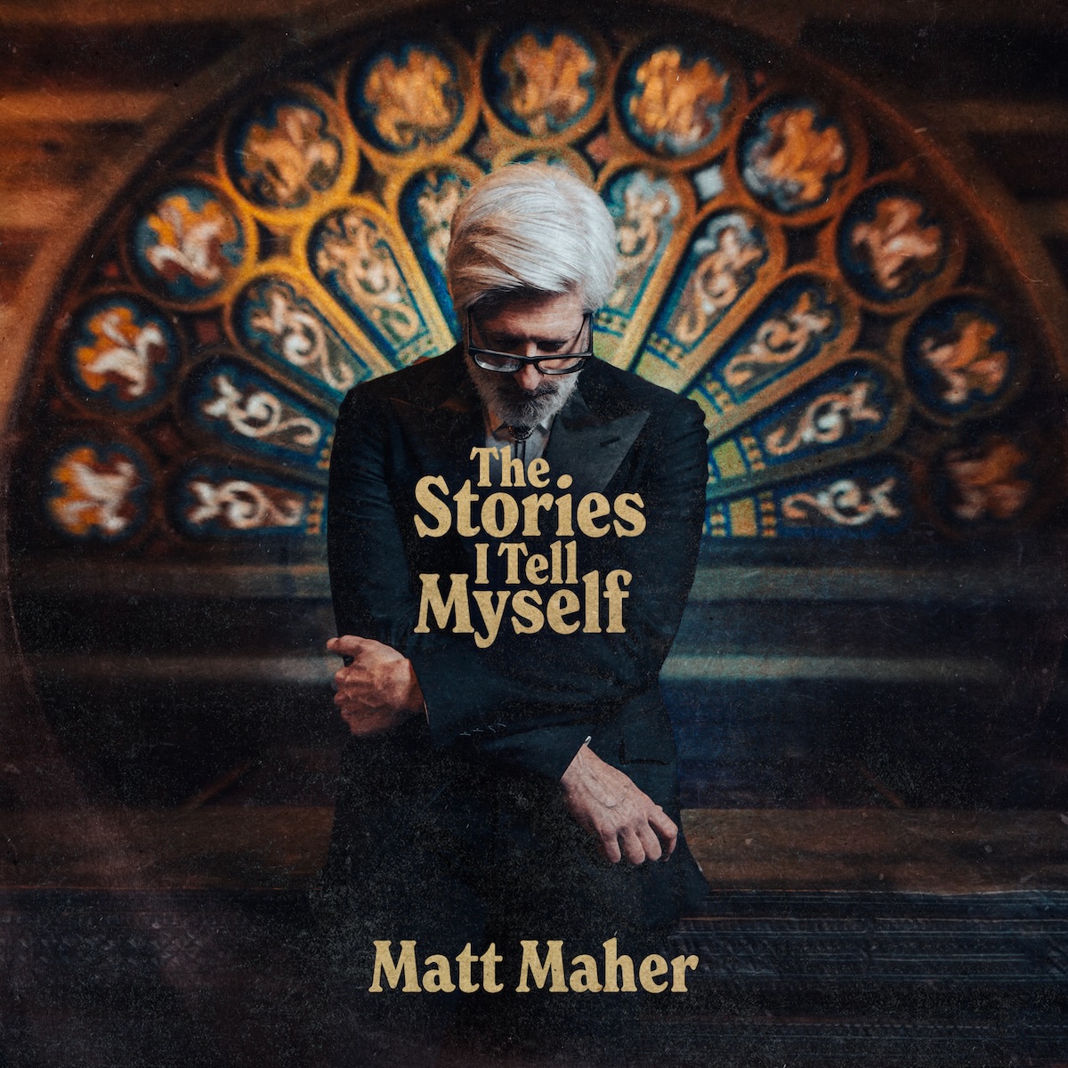 Album Art of Matt Maher's The Stories I Tell Myself album - A photo of a middle-aged man with white hair standing with his head bowed towards the camera and his arms crossed over his chest in a reflective or prayerful moment. He's wearing a black suit coat with a light-colored shirt and bolo tie. Behind him, artwork that looks like a half-circle rose stained glass window not lit from behind, but lit from the front and it still looks beautiful even if a bit out of focus.