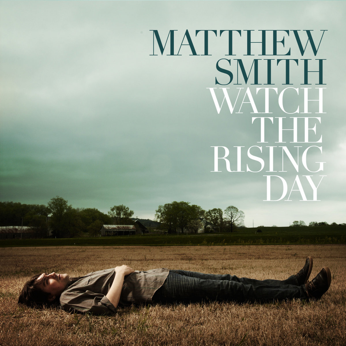 Album Art of Matthew Smith's Watch The Rising Day album - In a field of brown grasses, a photo of a man laying down in the grass and looking up into the green, cloudy sky is shown. In the background looks to be some trees and some farm buildings. and the top half is all cloudy sky. The person, a white man with fairly short hair, is wearing dark pants and a brown shirt and has right hand over his stomach.</body></html>