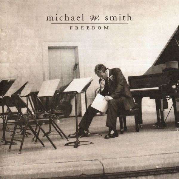 Album Art of Michael W. Smith's Freedom album - A black and white photo with a bit of a golden/brown sepia tone to it shows a lone man sitting on a stool in front of a grand piano, but facing away from the piano towards a small row of empty chairs with music stands and music sheets on the stands. The man is hunched over in contemplation holding the music sheets in his left hand, with his right hand up to his lips and looking down. The background is a wall with a door in the middle, and the stone floor comes to an edge at the front indicating it may be some sort of stage he is sitting on alone. He is middle-aged and wearing a full suit coat and pants.