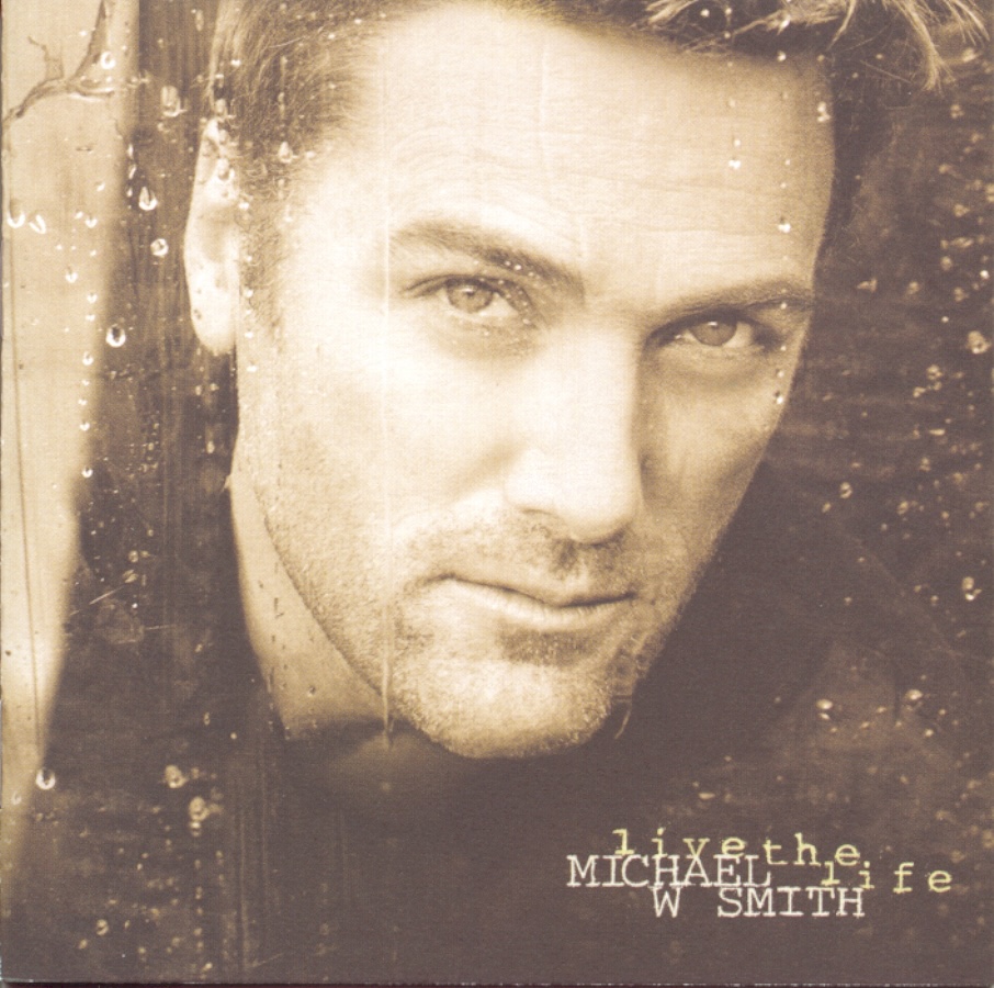 Album Art of Michael W. Smith's Live The Life album - A sepia-tone brown photo of a white man's face, looking through a window with raindrops on it. The man is looking at the camera and has a hint of a smile and his hair is short, and his beard and mustache are very short, mostly just a bit of fuzz. In the bottom right, in front of a dark brown background, is the artist's name in a white uppercase typewriter font, with the album title in lowercase golden brown to the right and above his name.
