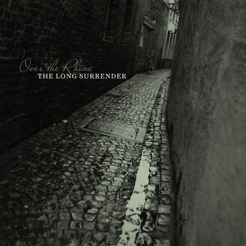 Album Art of Over The Rhine's The Long Surrender album - A dark, blank-and-white photo of a small, cobbled alleyway or narrow street in some sort of old city. As the dark street rounds a corner, a bike is just propped against the wall. Most of the picture is taken up with the walls on both sides of the alleyway and the stones of the street which are rough and wet. Only far-off down the alley can we get a glimpse of some buildings that are not just a blank wall.</body></html>