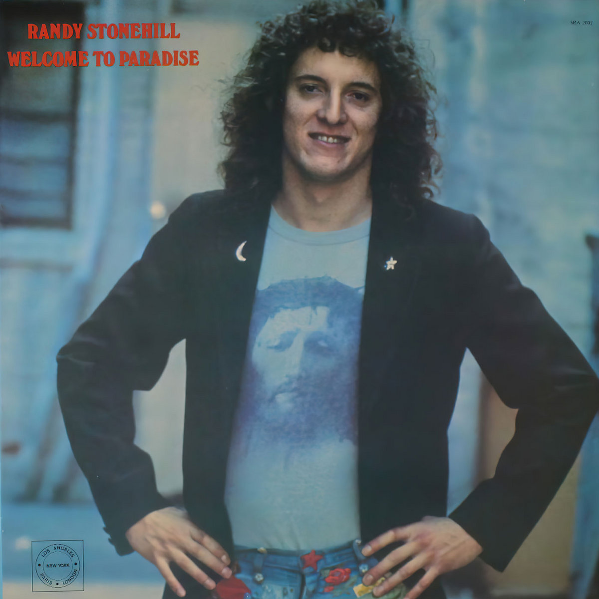 Album Art of Randy Stonehill's Welcome To Paradise album - A photo of a young man from the waist up. He's wearing blue jeans with colorful patches on them, then a T-shirt with a print of Jesus on them. Over his arms he's got a dark blue coat with pins of the moon on the left side of his chest and a star on the right side. His hair is dark, curly and rather large, a bit of a fro. The background seems to be just an outdoor scene with a house or other building behind him. In the top left, in red letters, is printed the artist name and the album title.