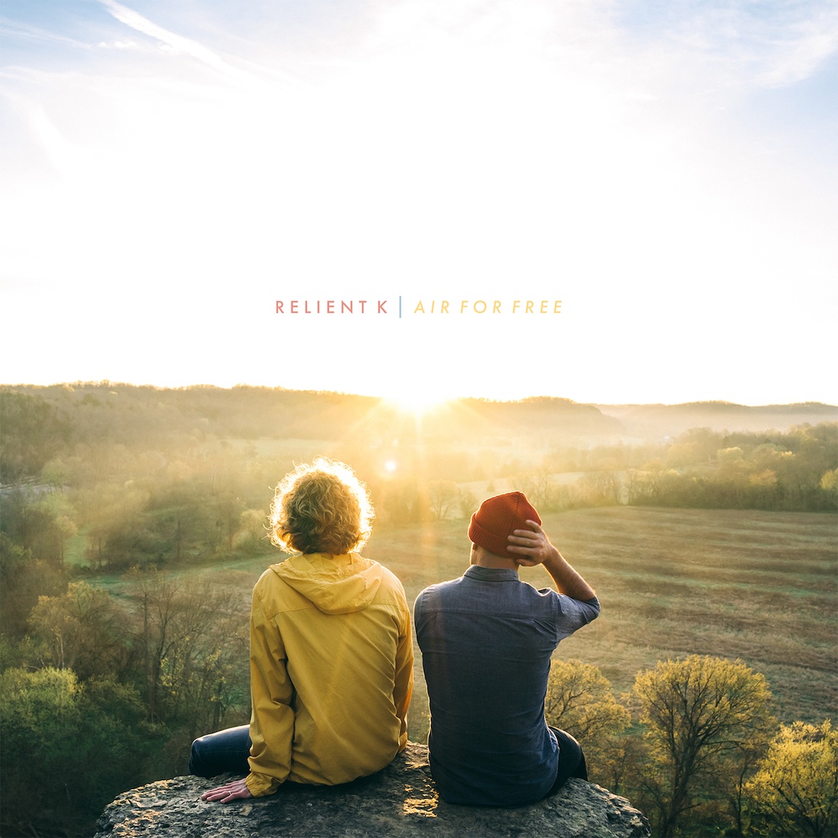Album Art of Relient K's Air For Free album - Sitting on a rock overlooking some fields with some occasional trees and maybe a forest and hills off in the distance, two men are looking towards the sun which is low on the horizon with their backs turned to the camera. The sun is bright and the brightness makes most of the partly cloudy sky white and the landscape a bit hard to see. The man on the left has curly, light brown hair and is wearing a yellow hoodie and blue jeans. The man on the right is wearing a denim jacket and a red beanie and has his right hand adjusting his hat. Above where the sun is is printed the band name in uppercase red text, a vertical blue line, and then the album title in yellow text.