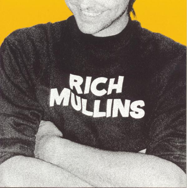 Album Art of Rich Mullins's self-titled album - A black and white photo of a man standing against a bright yellow background. only the bottom of his face, a grin, can be seen at the top. The center of the photo is is chest with his armed crossed below, and the black T-shirt with 'Rich Mullins' written in large, white text in the middle of the shirt. The photo is pretty grainy.