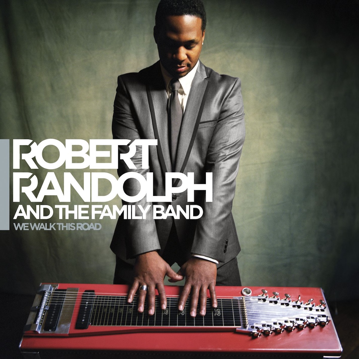 Album Art of Robert Randolph & The Family Band's We Walk This Road album - A photo of a young, black man in a very nice metallic gray suit is standing in front of a slide guitar with a bright red housing, and his hands are just touching the strings. He's looking down at the instrument and the background seems to be a pretty standard photo studio backdrop of a marble pattern lit more in the middle. Above the photo is printed the artist's name in large white sans-serif font off to the left, with 'and the family band' below it smaller. Even smaller below that is the album title in gray.