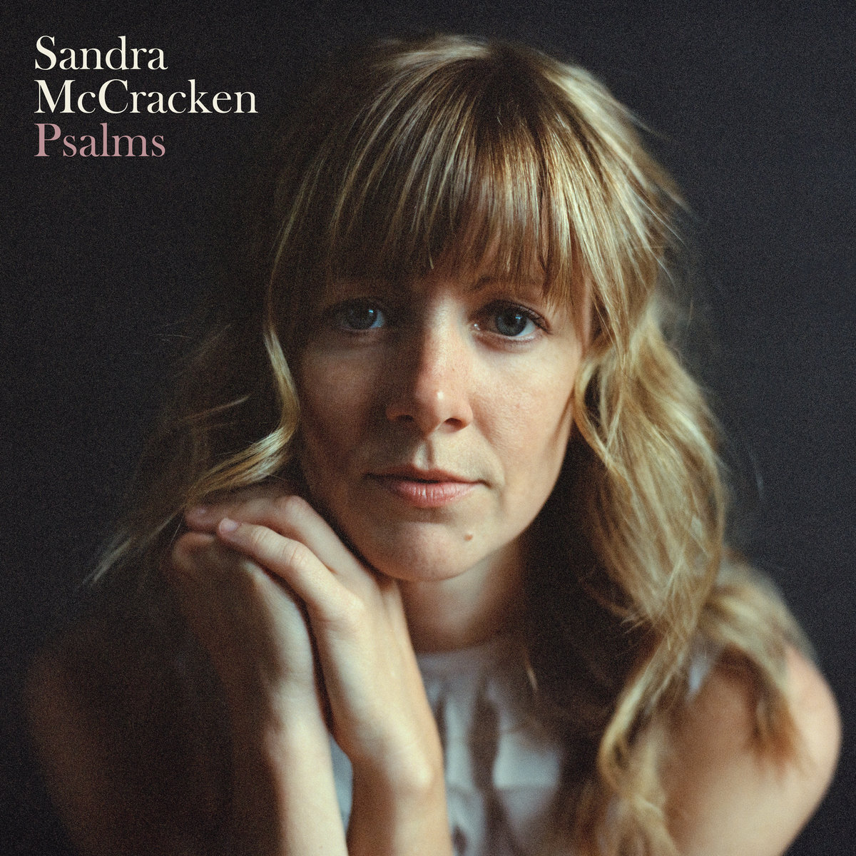 Album Art of Sandra McCracken's Psalms album - A close-up photo of a middle-aged woman from the shoulders up, with her hands clasped together to the left of her chin. Her blone-brown hair goes down past her shoulders. She is leaning in and staring straight into the camera with a fairly blank expression. The shoulders are out of focus. The background is a dark gray, nearly black. In the top left, printed in a serif font is the artist name on two lines in a milky white, with the album name just below it in a pastel pink.