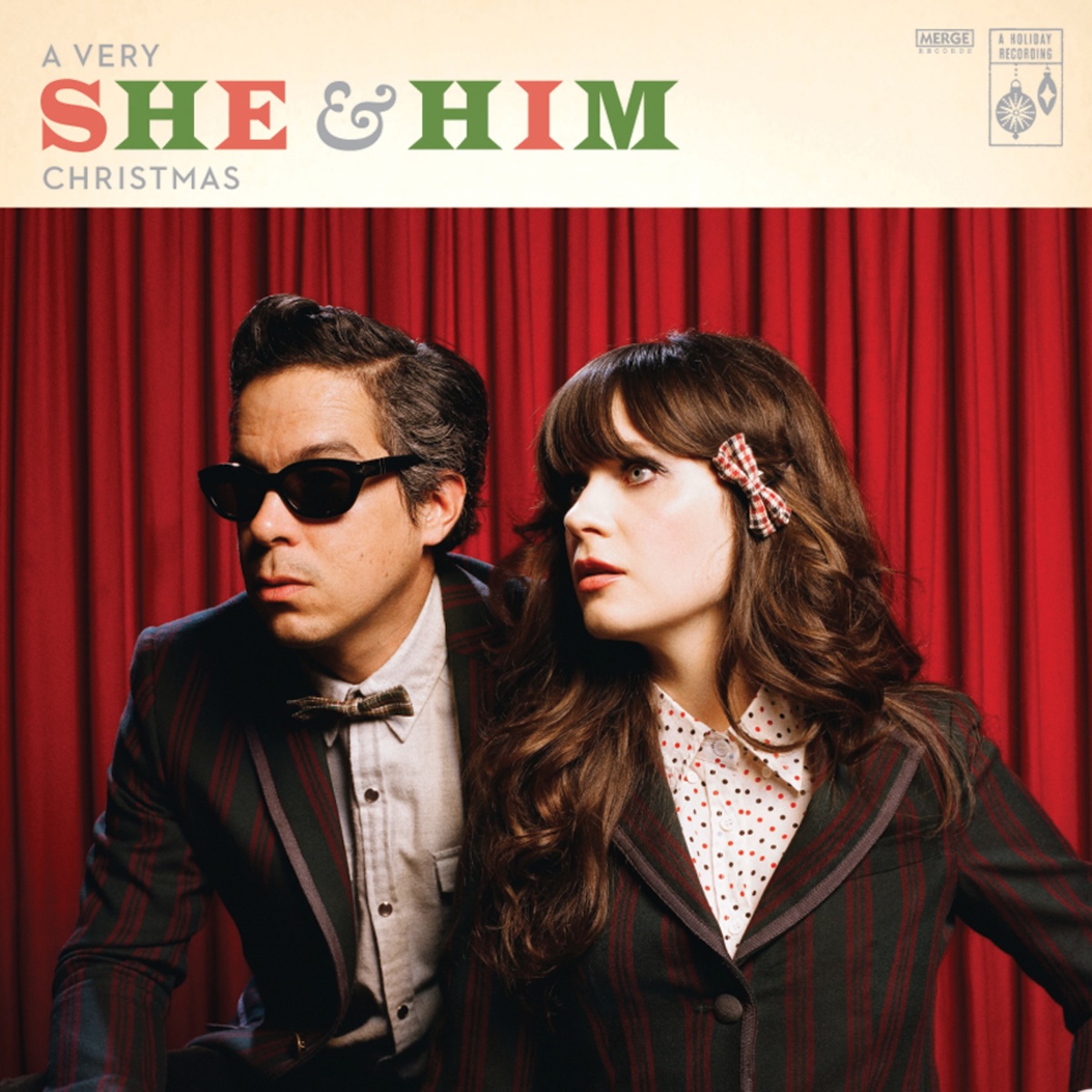 Album Art of She And Him's A Very She And Him Christmas album - The bottom four fifths is a photo of a white man and white woman from the chest up wearing black suits and white-colored shirts beneath. They are in front of a red curtain and seem to be looking at something to the left of frame with some awe or surprise. The man has a bowtie and is wearing sunglasses.</body></html>