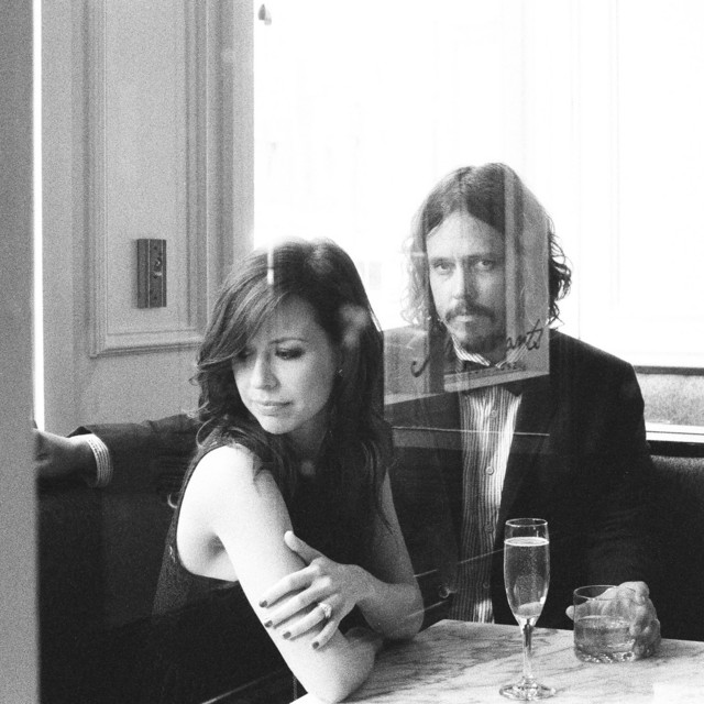 Album Art of The Civil Wars's Barton Hollow album - A black and white photo of two young white people, on the right a man with curly hair, a beard and mustache in a suit coat with a bowtie, and on the left, a woman with darker, long hair and a sleeveless dark top. They are sitting in a corner booth with a marble tabletop sitting in front of them and glasses of water on the table. The man is looking at the camera and has his right arm behind the woman and the woman is looking away from the man to the left. There is lots of light behind them as the area has many windows, it seems. It seems like they also may be photographed through a window, because there's a reflection of another window overlaid in front of the couple.