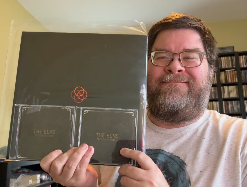 A photo of a man smiling on the right in a white-gray T-shirt with a green design holding a black vinyl album with four colored circles on the cover in the middle, plus two copies of the CD, with the black cover described separately in the other image.