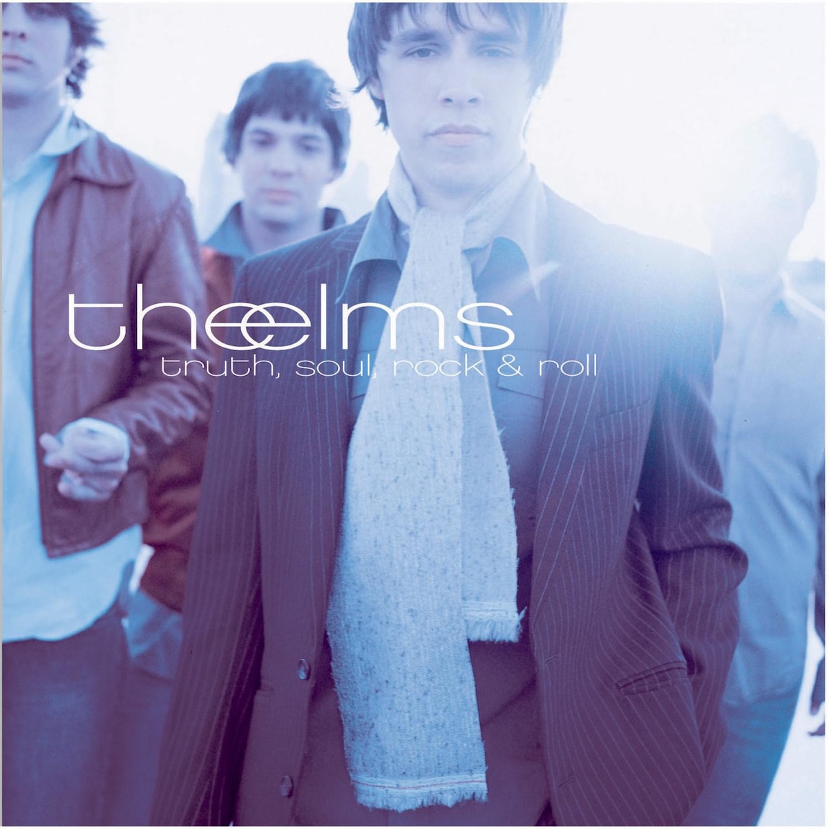 Album Art of The Elms's Truth, Soul, Rock And Roll album - A photo of four young men in collared shirts and coats. The one in the front has a pinstriped suit coat and a scarf. The man on the right's head is almost completely obscured by a bright light and is behind the one in the front, so not too much detail to make out of them there. They are all sporting short hair and their outfits all seem to have versions of dark red or blue colors. On top of the photo, vertically centered but on the left horizontally is the band's name in loercase, with the 'e' from 'the' and the 'e' from 'elms' overlapping. The text is all white and below it is the album title in a much smaller font.