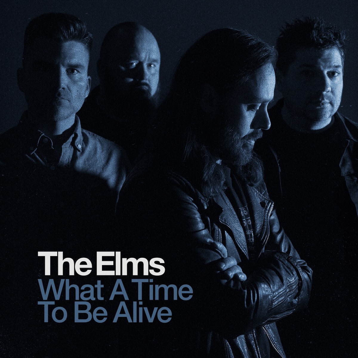 Album Art of The Elms's What A Time To Be Alive single - A photo of four middle-aged men in a black room, which soft blue lighting illuminating them from the right side of the frame. They all have at least stubble on their face or full beards. Three are looking at the camera, while one is looking down and off to the right. In the bottom right quarter, it has the band's name printed in white with the album title in a dark blue spread over two lines below it.