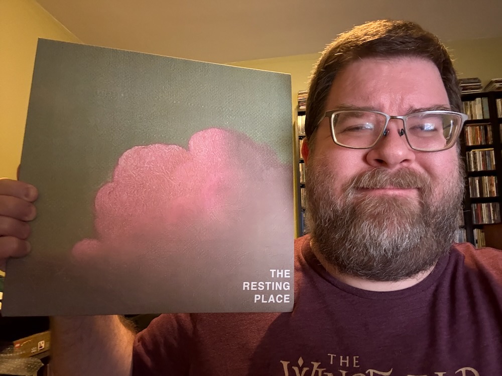 Photo of a middle-aged man from the chest up on the right, holding a vinyl album in a slipcase on his left. The album is The Resting Place's self-titled album. He's got a beard, a mustache, and thick glasses. Behind him is a shelf with many CDs and the walls are a light yellow.