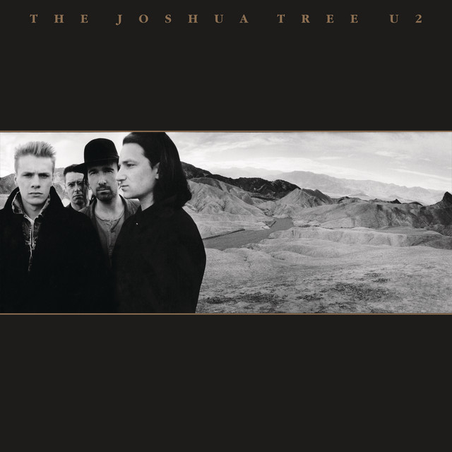 Album Art of U2's The Joshua Tree album - The majority is a black background on the top and bottom. At the very top, the album title and band name are printed in small, gold letters. In the middle third, surrounded by a gold line at the top and bottom, is a black and white photo of four men. The left 3 are looking a the camera, and the fourth is looking of to the left. They're all wearing dark jackets, and the third one has a dark bowler hat. To the right of the band, a rocky terrain is shown and stretches off to mountains in the distance.