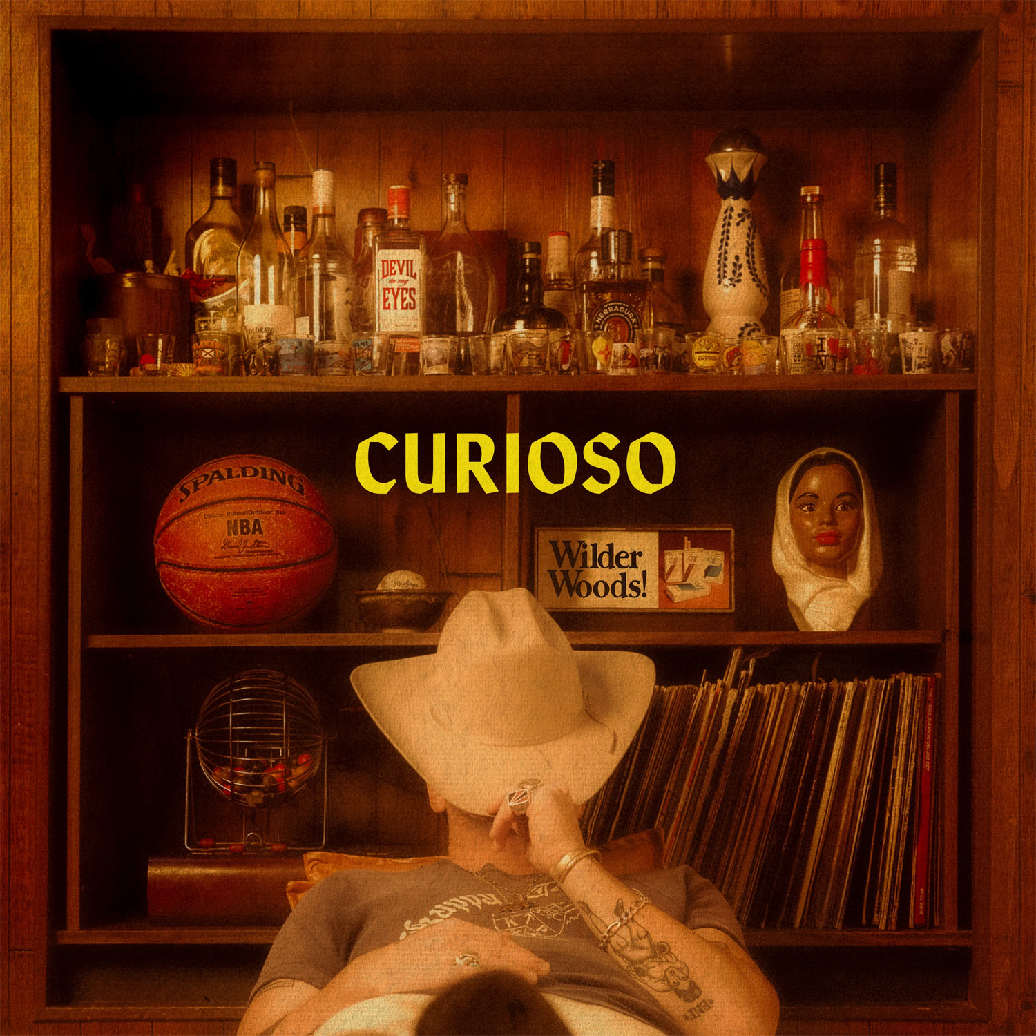 Album Art of Wilder Woods's Curioso album - A photo of a man sitting on a nice chair in front of a bookshelf, though there's no books here. The top shelf is dozens of liquor bottles and dozens of shot glasses. The middle shelf has an NBA basketball, a baseball in a bowl, and cigarette ad that says 'Wilder Woods!', plus a bust that looks like a dark-skinned woman with a head covering. On the last shelf, there's about 30 records, maybe. The man is leaning back in a chair, wearing a T-shirt, and has a cowboy hat over his face. The man has a number of large rings on, a golden armband, and a number of tattoos on his arms. In the middle, the word 'Curioso' is printed in bright yellow.